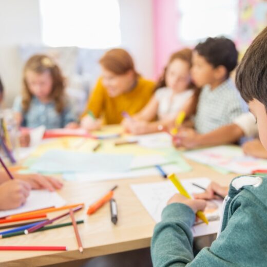 Children sitting at a table, focused on drawing with colored pencils and markers on paper. An adult is assisting in the background.