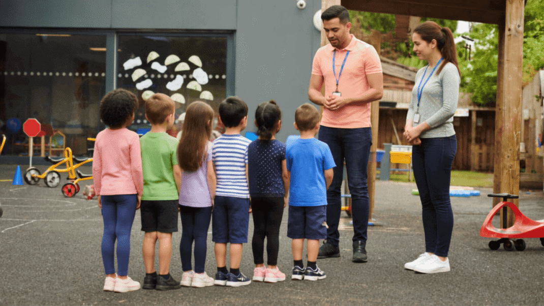 A male and female teacher stand facing a small group of children in a school playground.